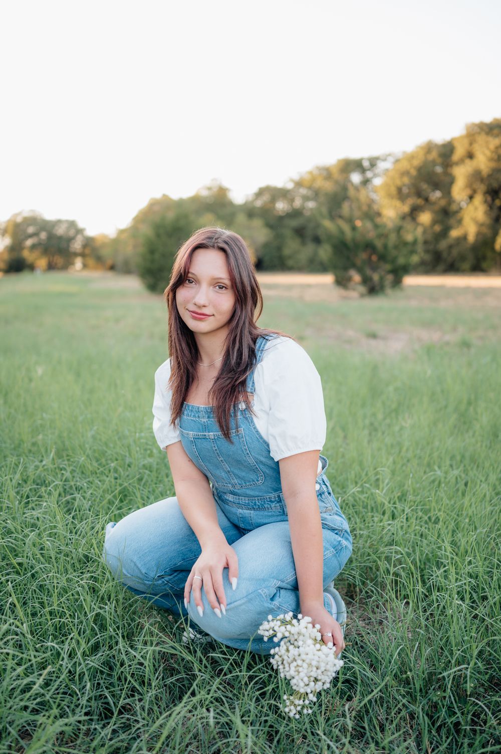 Person in denim overalls sits in grassy field holding wildflowers at sunset.
