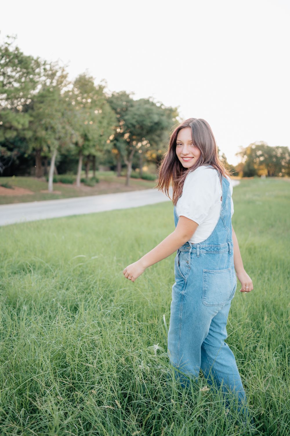 Person in denim overalls twirls playfully in grassy field during sunset.