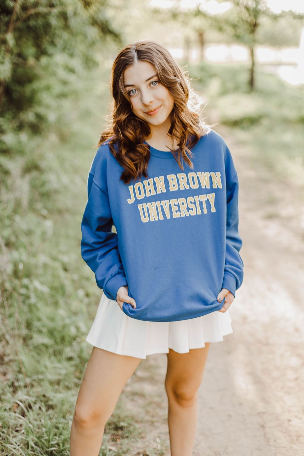 Student poses in John Brown University sweatshirt and white skirt on campus path.