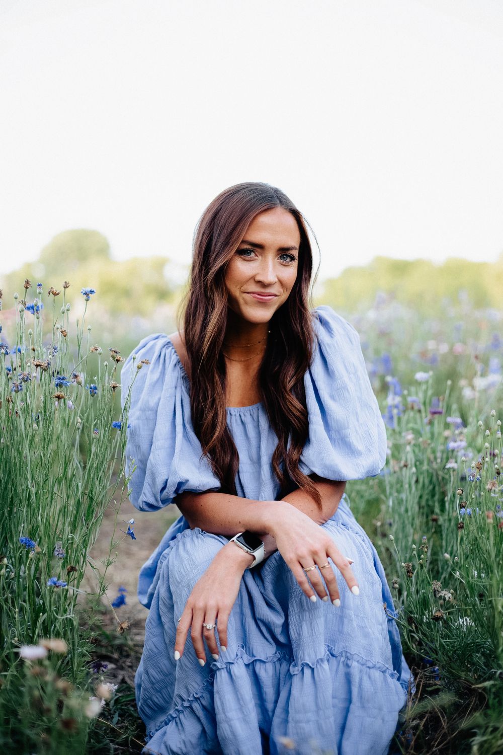 Woman in blue prairie dress sits in field of wildflowers during sunset photo session.