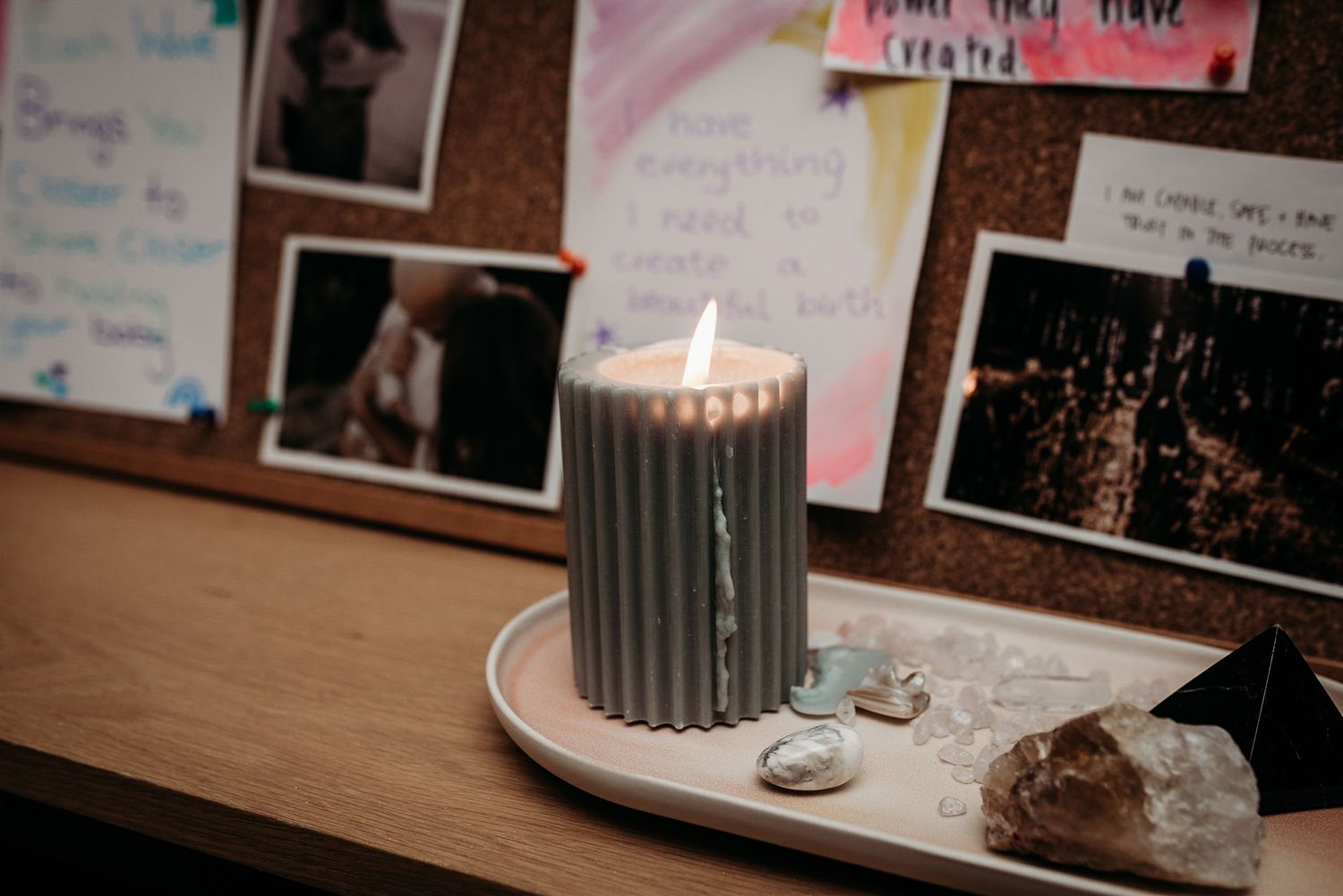 Gray candle burning on a plate with crystals and photos on a cork board in background.