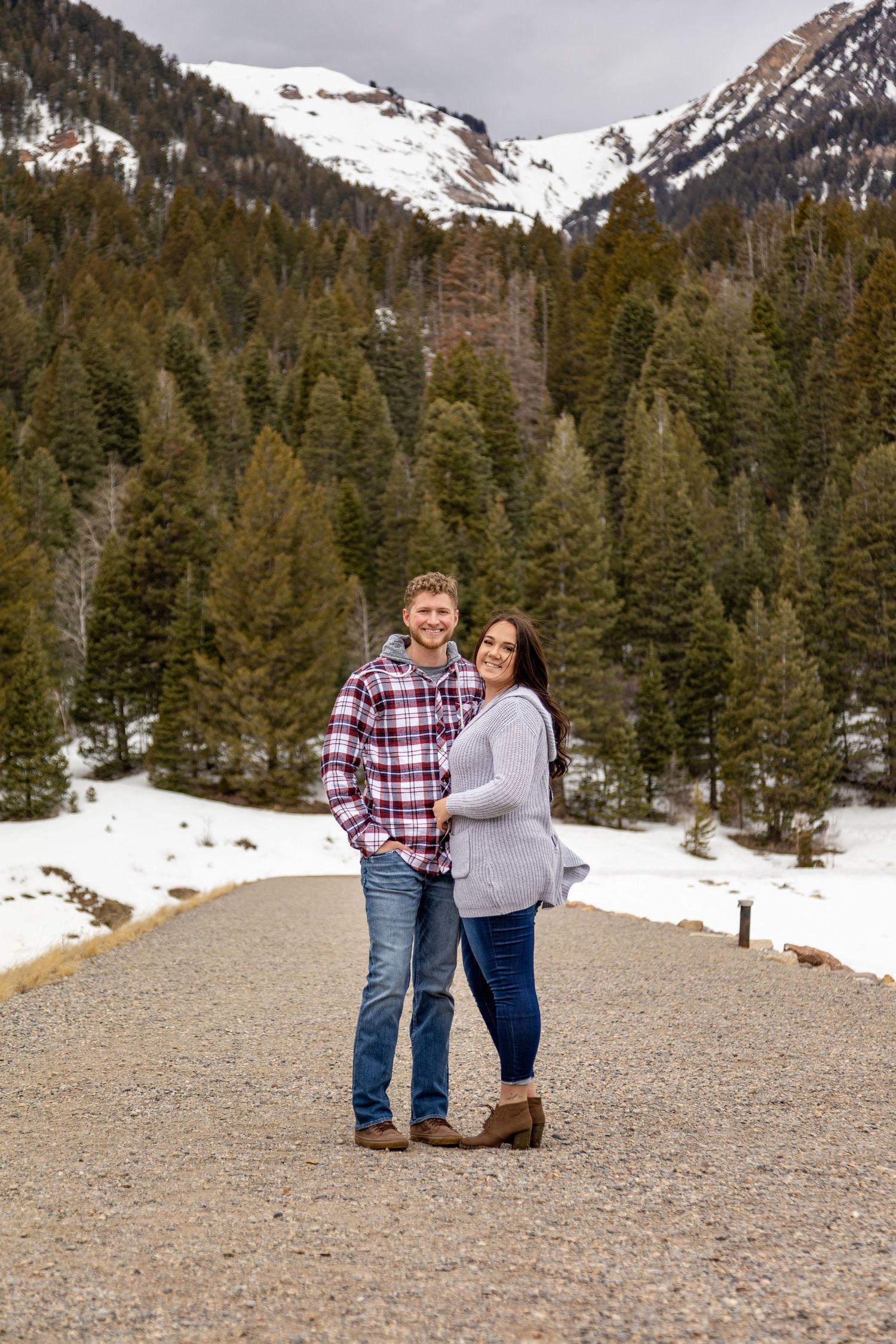 A couple stands together on a gravel path with snow-capped mountains in the background.