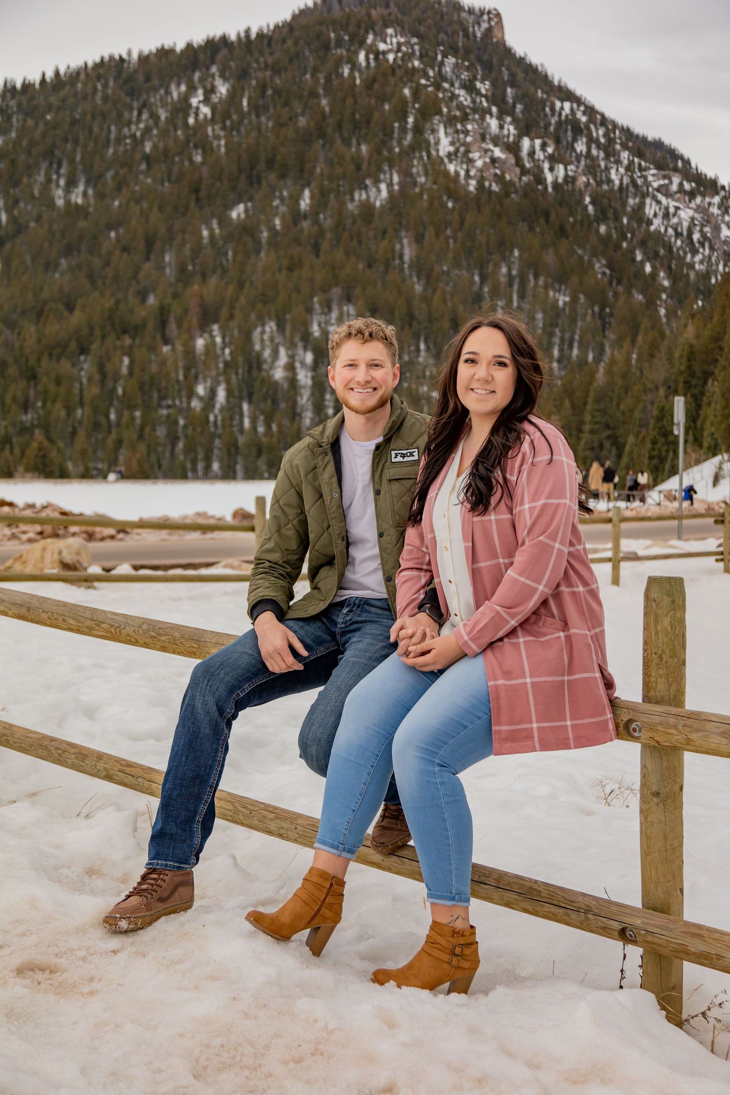 A couple sits on a wooden fence with snow-covered mountains and pine trees in the background.