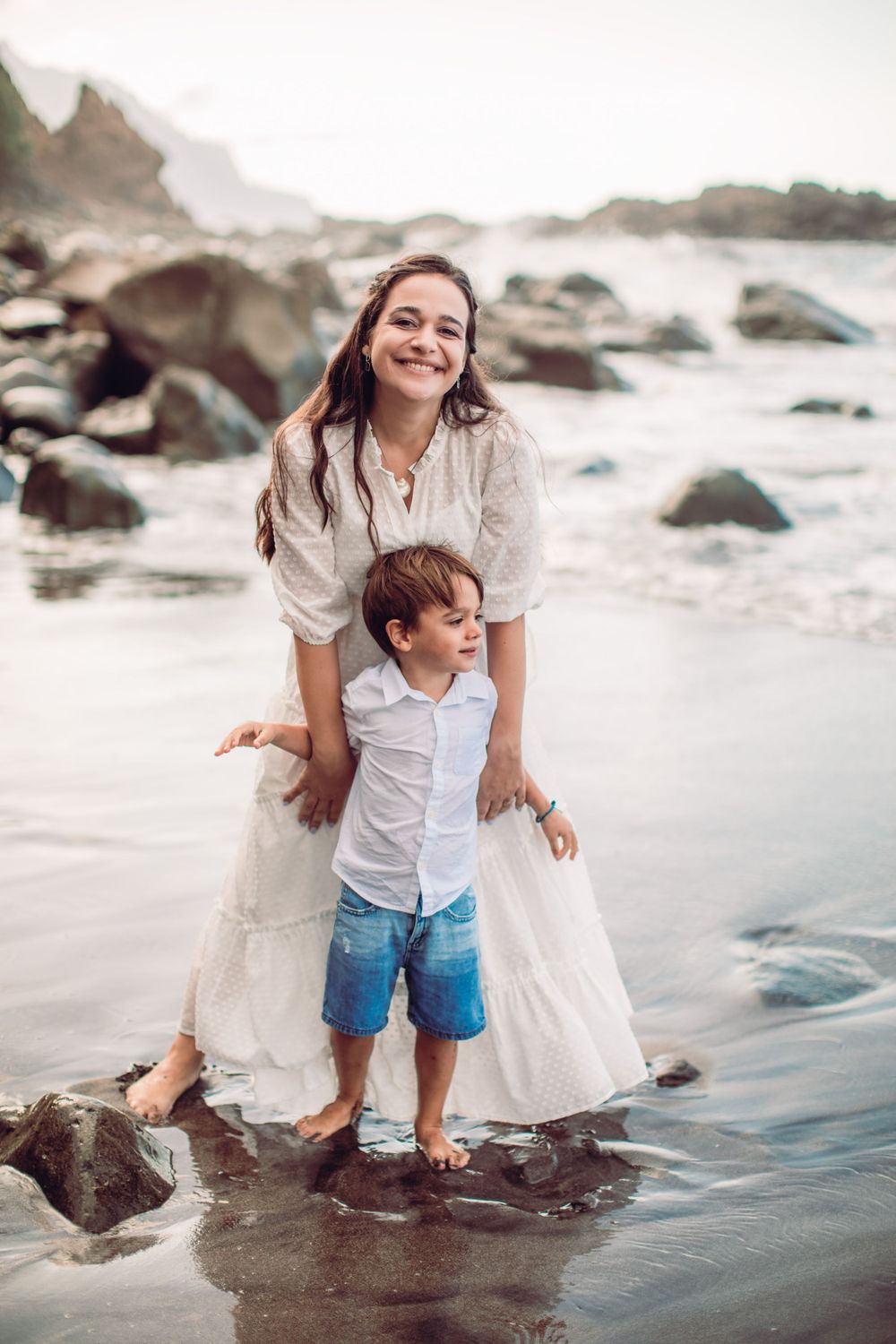 Parent and child playing in shallow ocean water near rocky coastline.