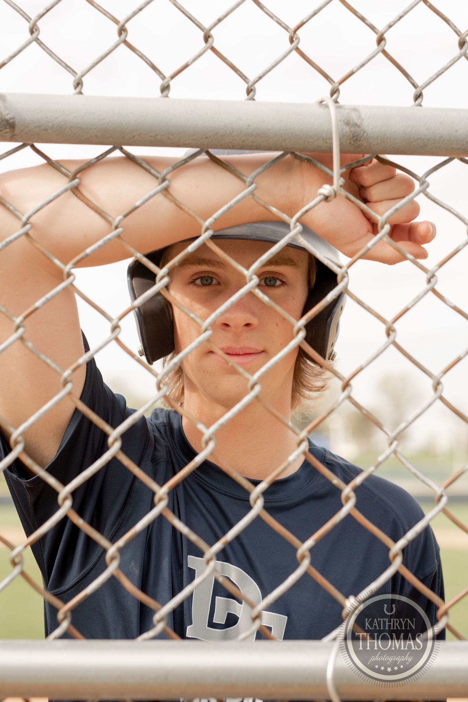 Baseball player looks through chain-link fence wearing navy uniform and helmet.