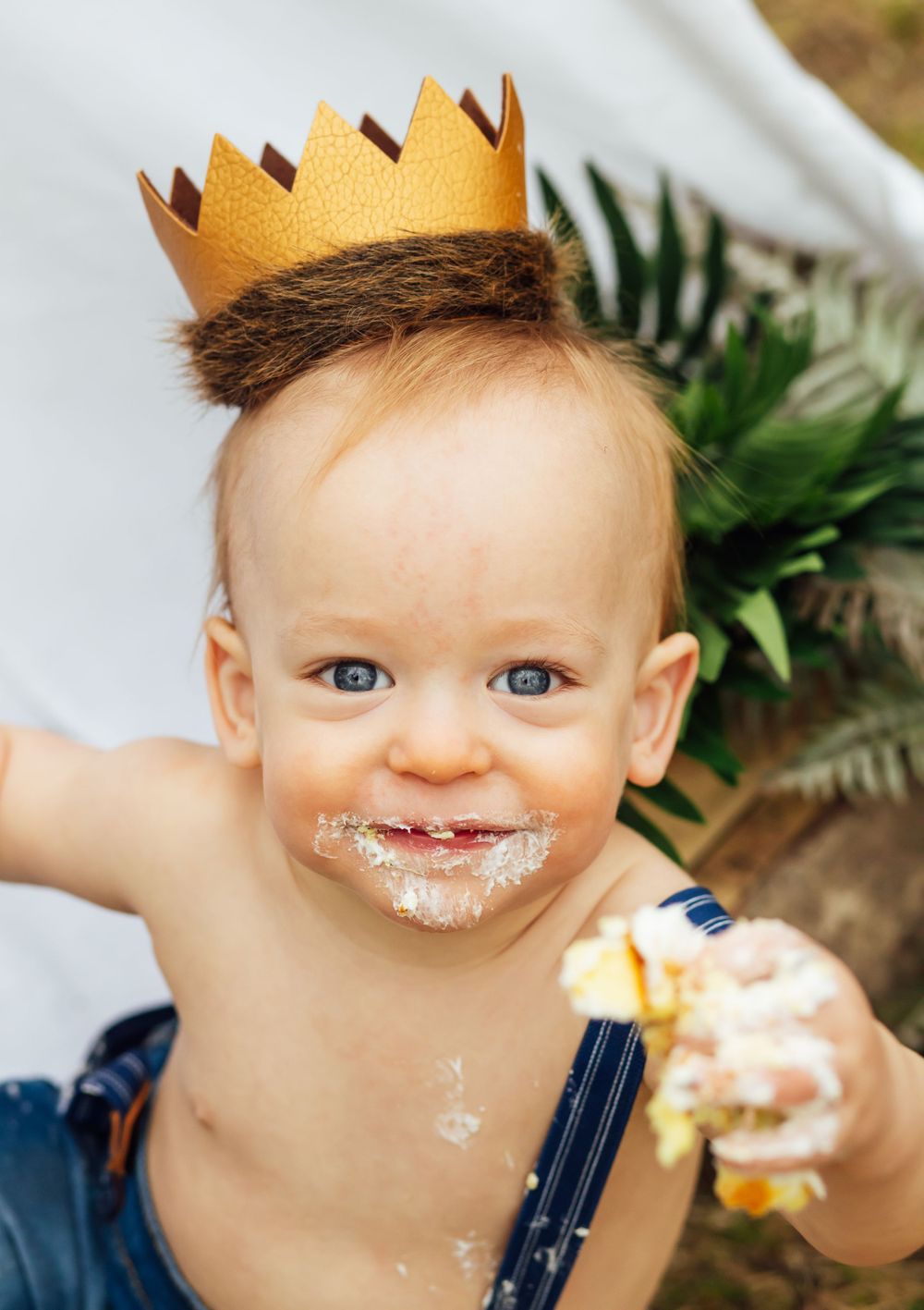 A small child wearing a gold paper crown eats cake with a messy, happy expression.