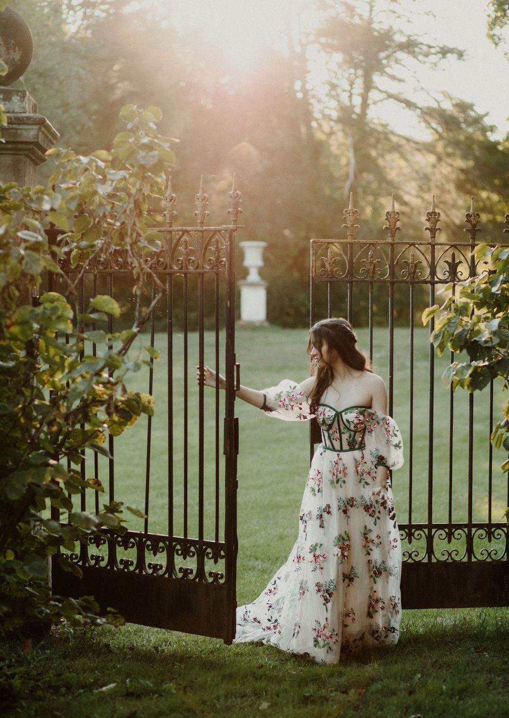 A woman in a floral dress stands at an ornate garden gate during golden hour.