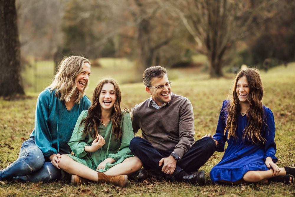 A family group sits together laughing on grass in an autumn field.