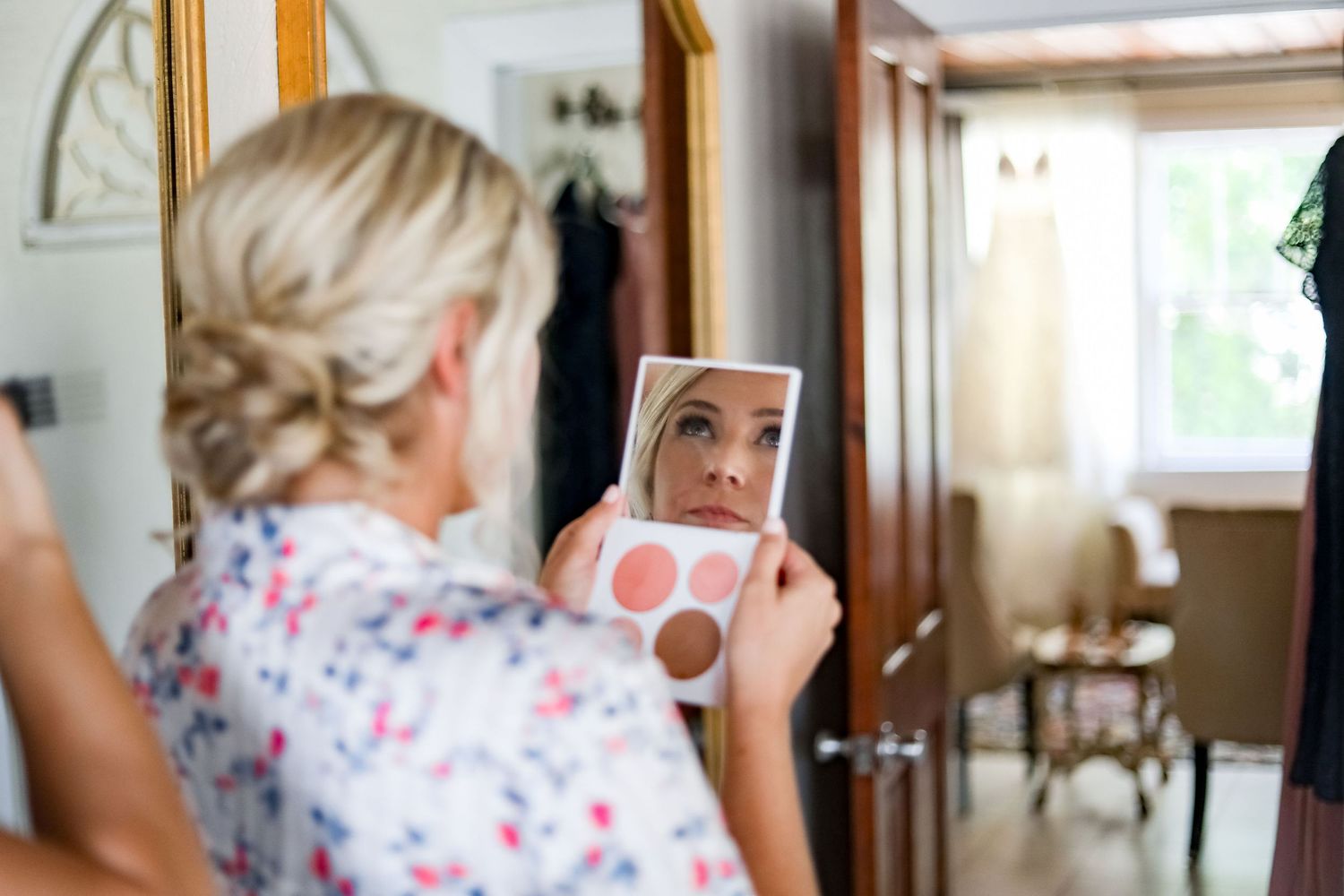 Someone applies makeup while looking in a small hand mirror wearing a floral print blouse in natural light.