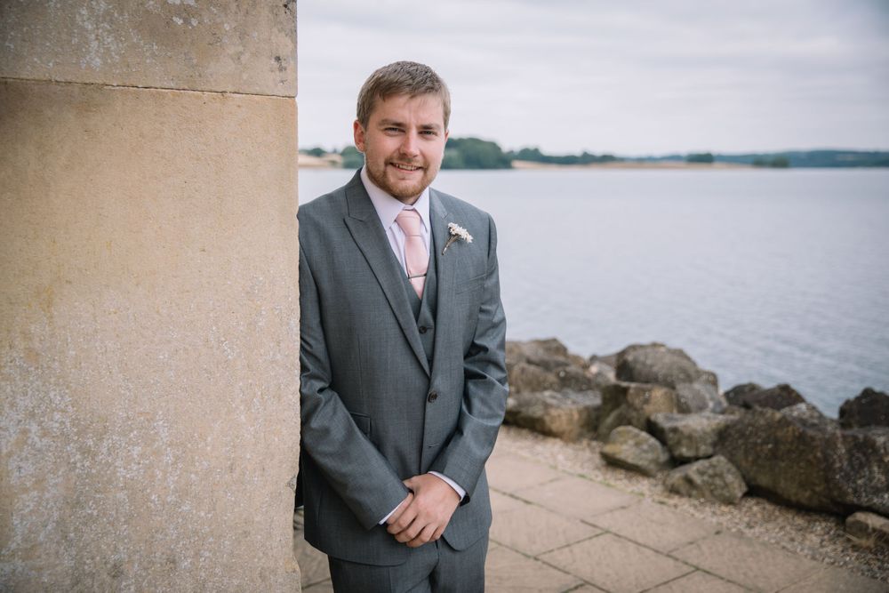 Groom in grey suit stands beside stone pillar with waterfront view in background.
