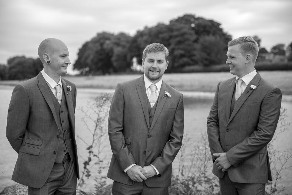 Three groomsmen in matching gray suits share a candid moment outdoors in a black and white photo.