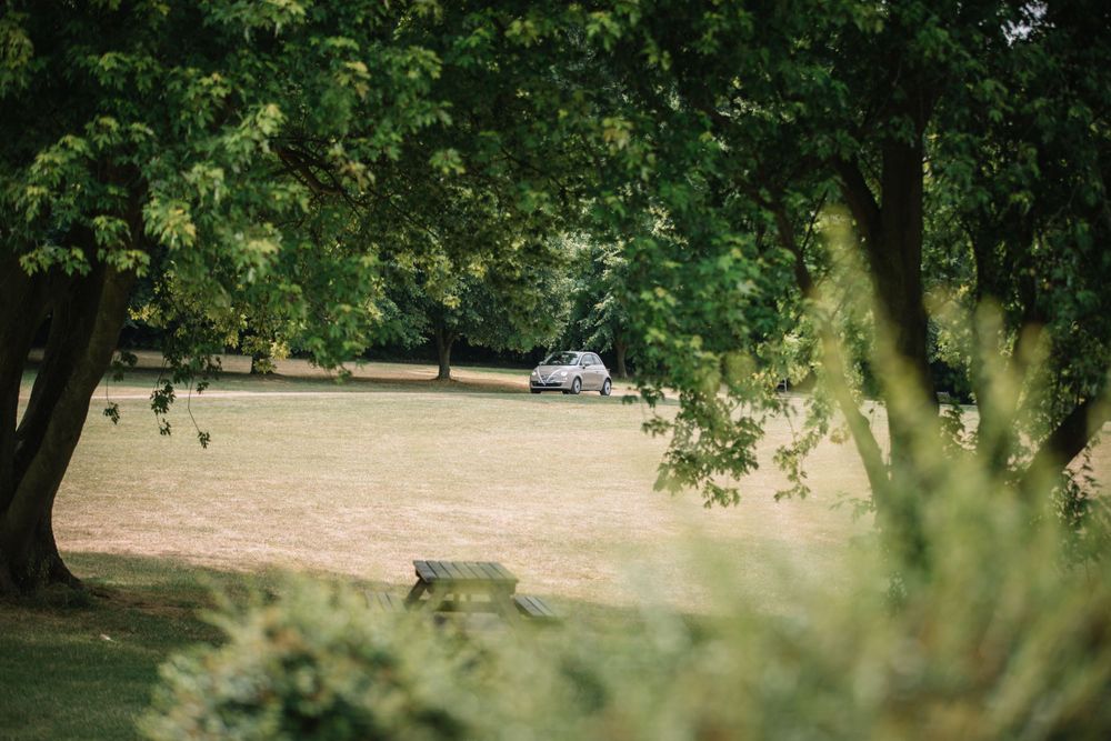A car is visible through trees and foliage in a peaceful park setting.
