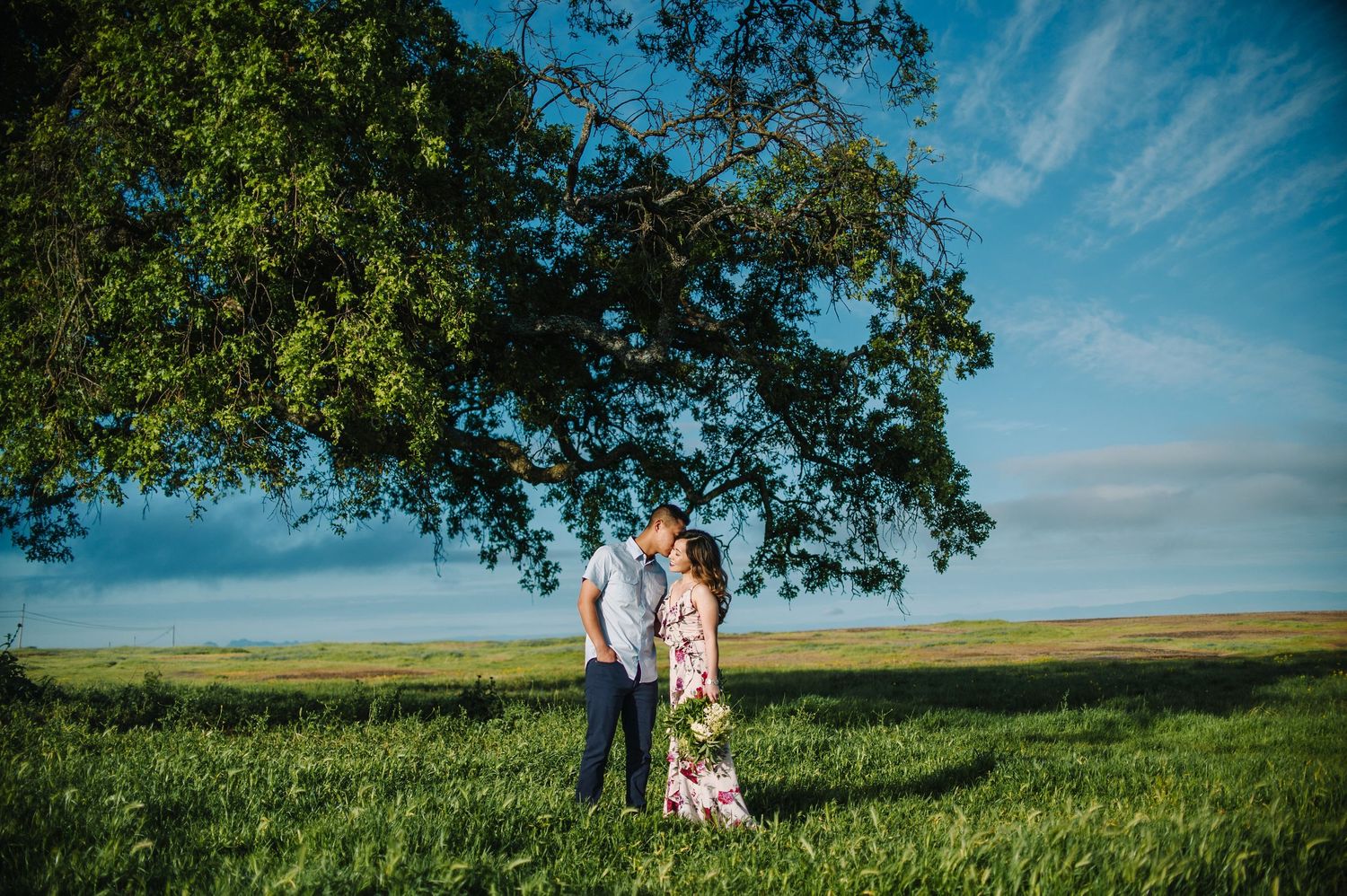 A couple stands under a large oak tree in a green field with their dog.