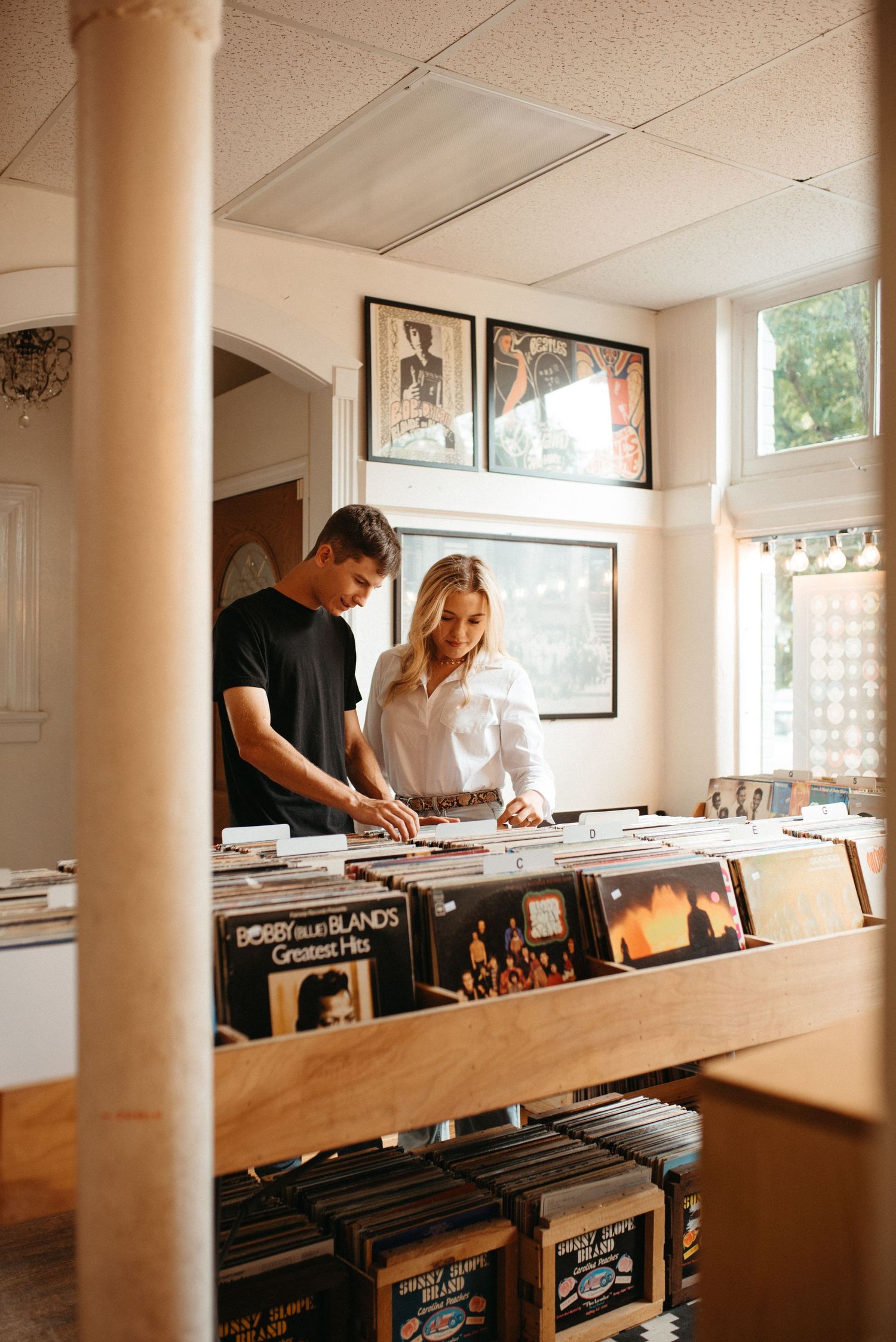 Shoppers browse vinyl records in a store with framed artwork on the walls and natural lighting.