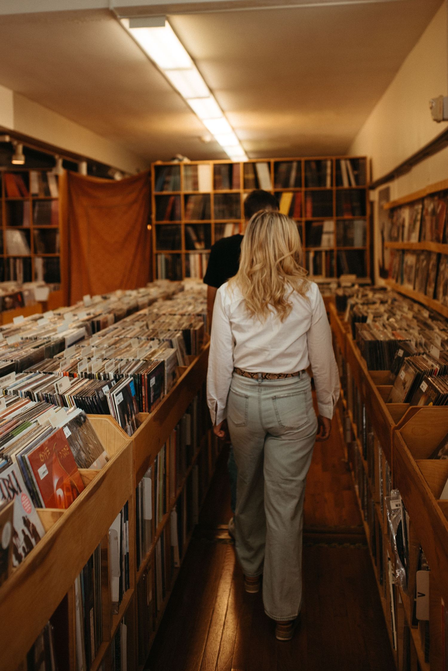 Person in white shirt walks down aisle of vintage record store with wooden bins and shelving.