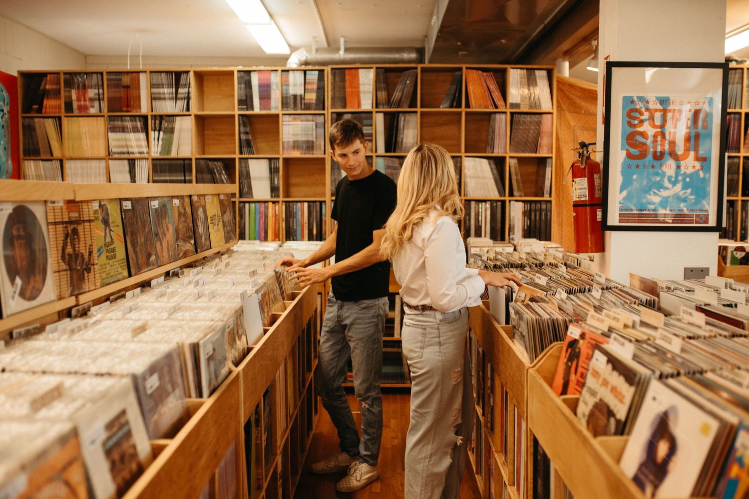 Shoppers explore vinyl selection in record store with wooden bins and wall-mounted album art.