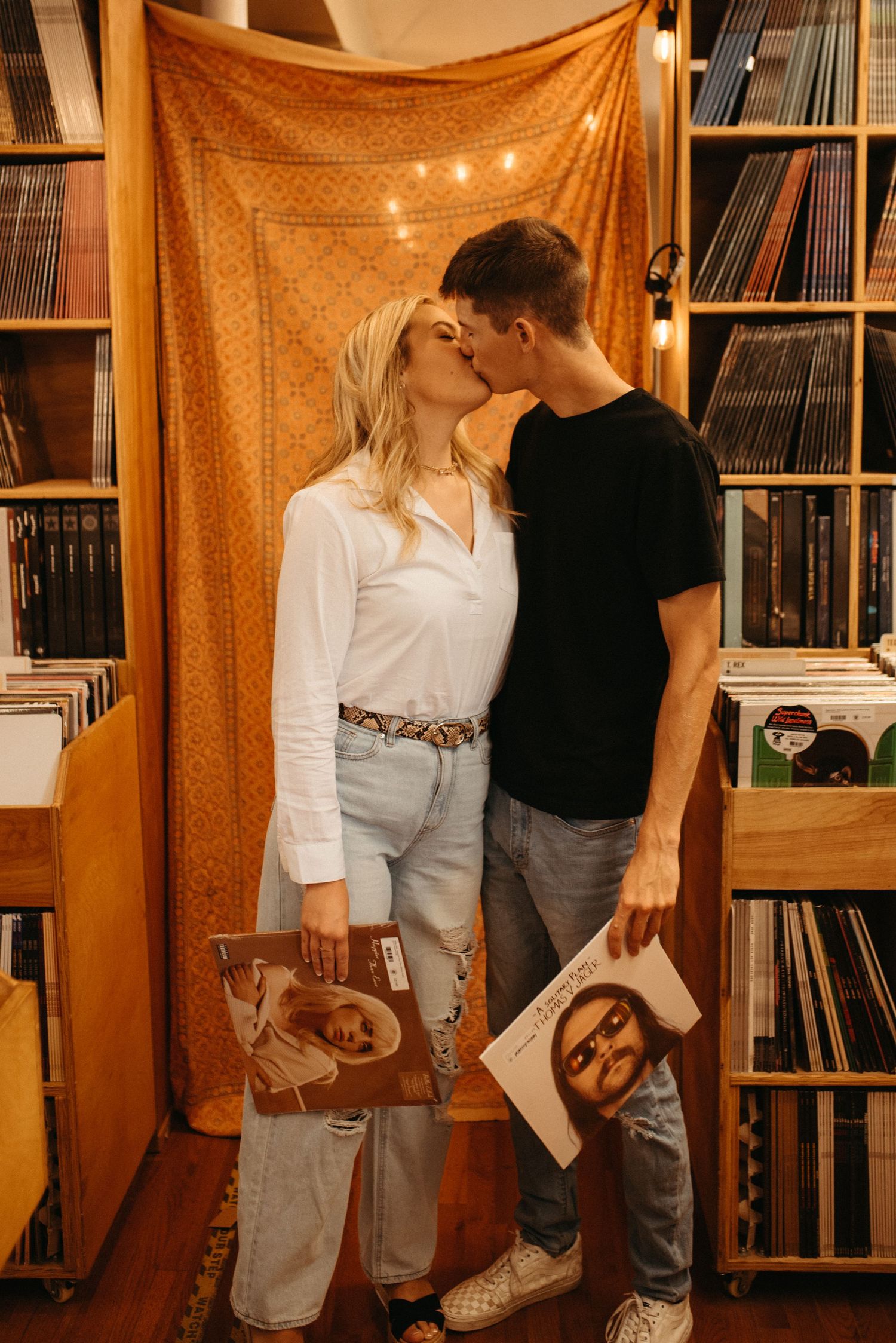 A romantic couple holding vinyl records while sharing a kiss in front of an orange tapestry.