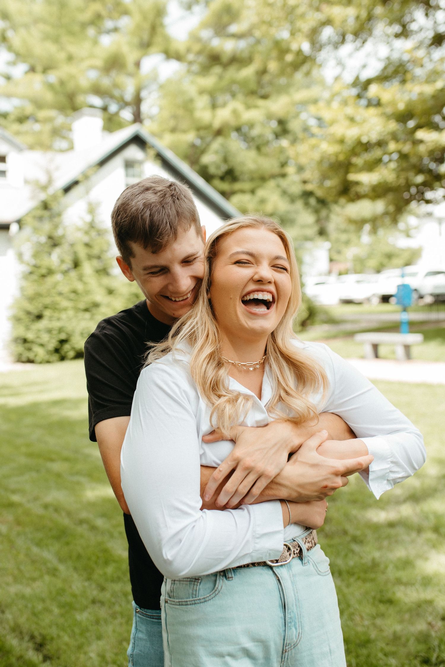 Joyful couple embracing and laughing in sunny backyard with white house in background.