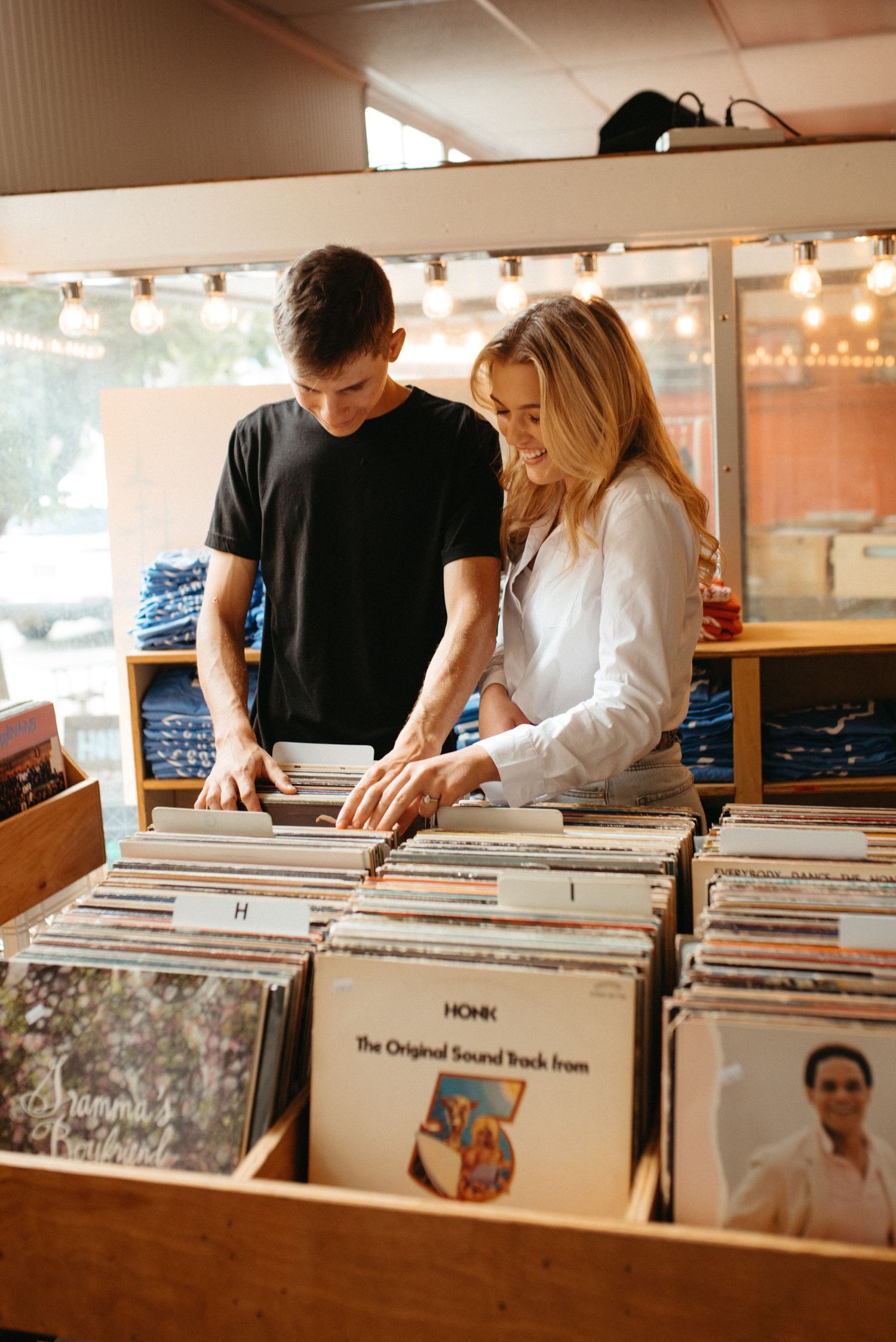 Two people flip through vinyl albums together at a record store with string lights in the background.