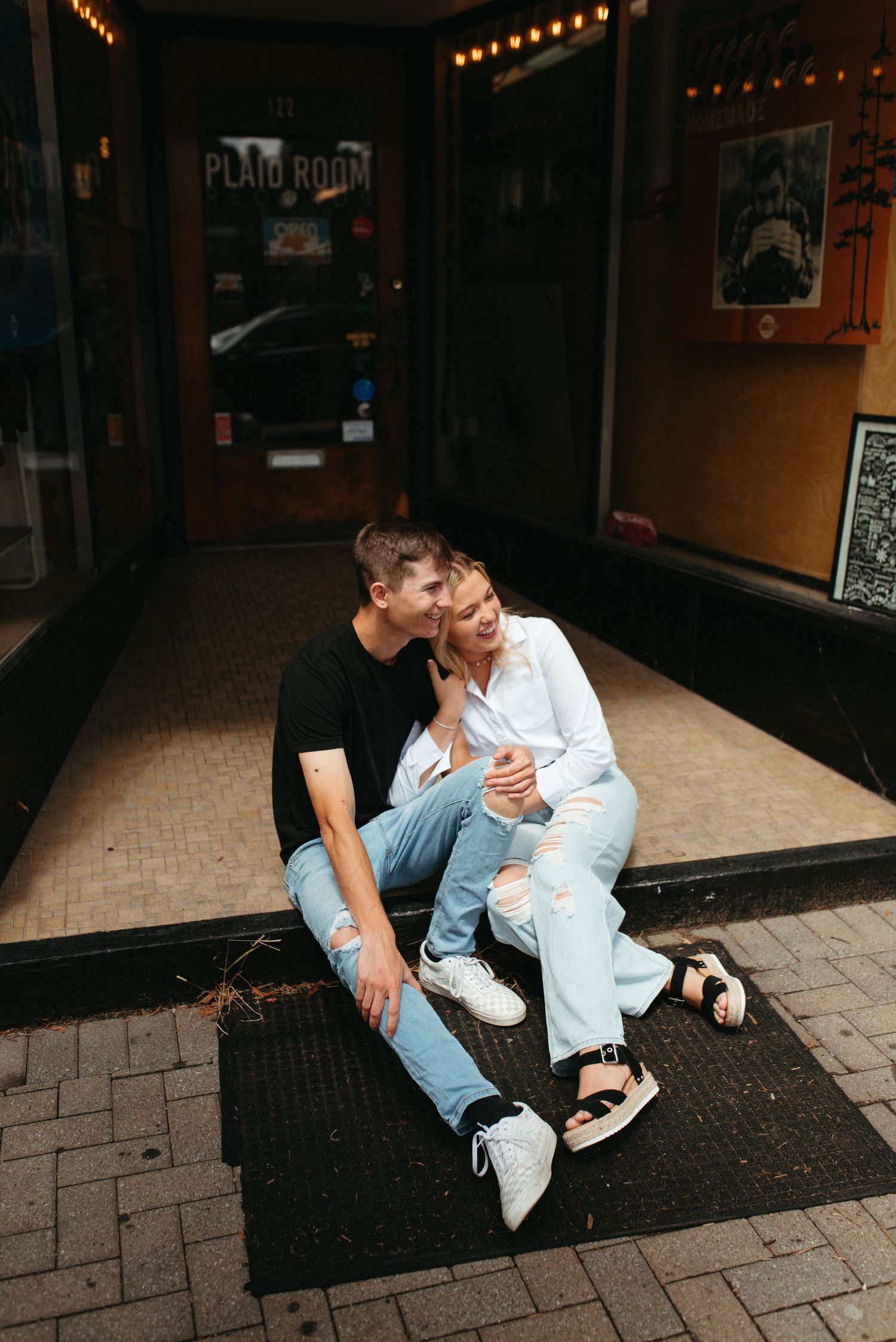 Couple in casual denim sitting together outside storefront with string lights.