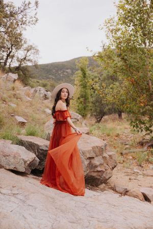 A flowing red dress catches the sunlight on rocky terrain with autumn trees in the background.
