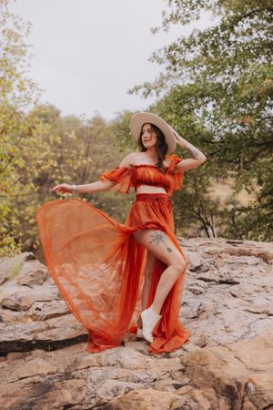 A flowing coral dress and wide-brimmed hat create a romantic silhouette against rocky landscape.