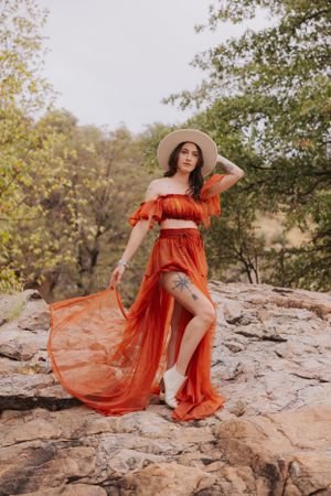 A dramatic orange dress with thigh-high slit flows in the breeze atop rocky terrain.