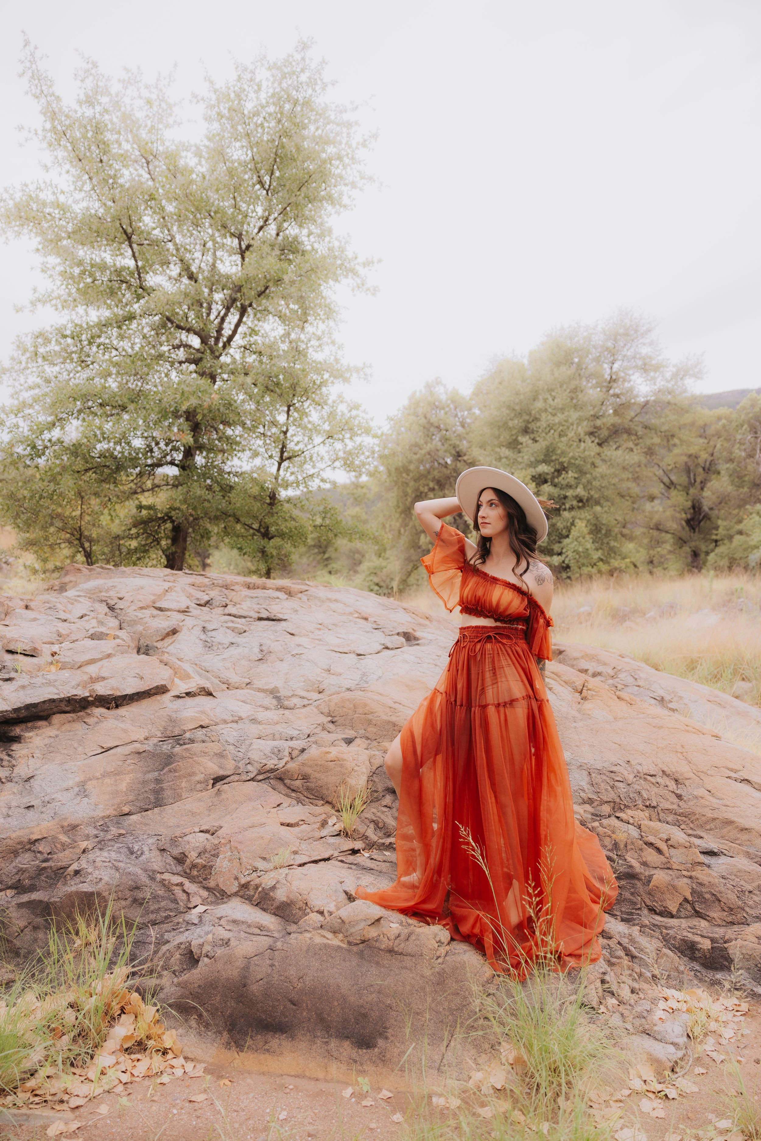 A flowing orange off-shoulder dress catches the wind as someone poses dramatically on rocky terrain.