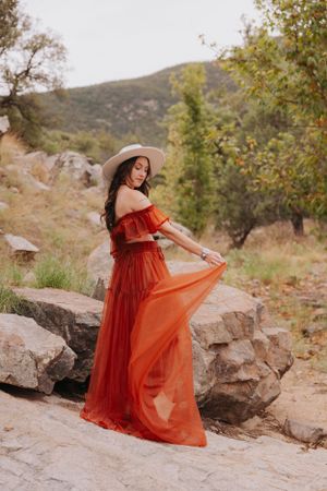 Dramatic red dress billows across rocky terrain with mountains and trees in the distance.