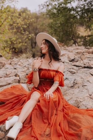 A rustic fashion shoot features a flowing orange dress and wide-brimmed hat on rocky terrain.