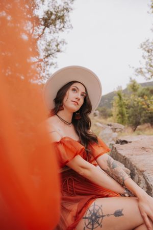 Artistic portrait with red dress and hat against natural mountain scenery.