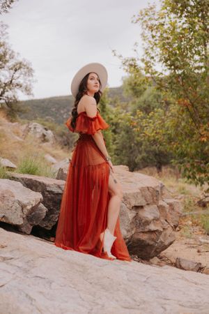 Wide-brimmed hat and red dress create romantic silhouette against mountain landscape.