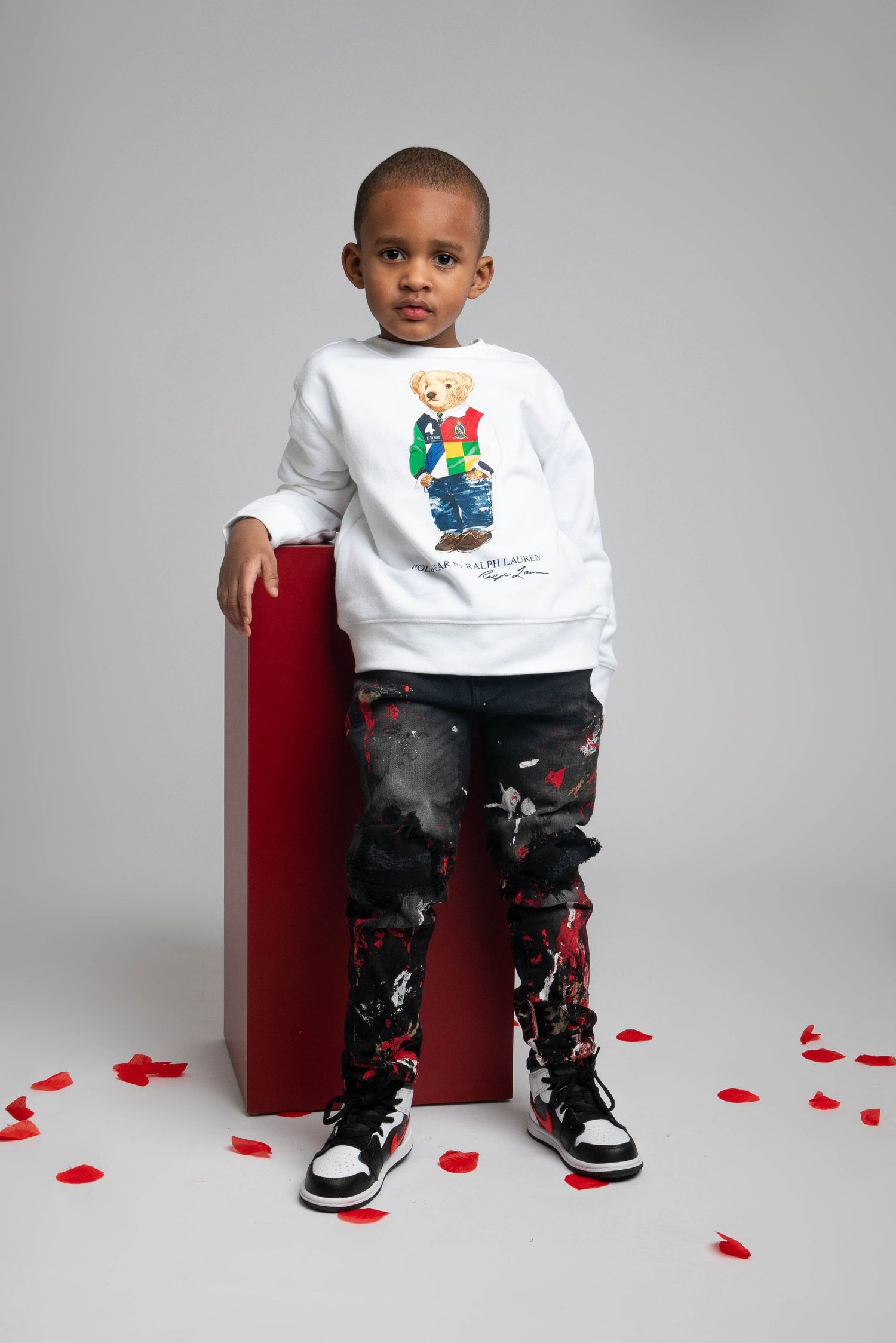 Child wearing white graphic sweatshirt and patterned pants on red bench against grey backdrop.