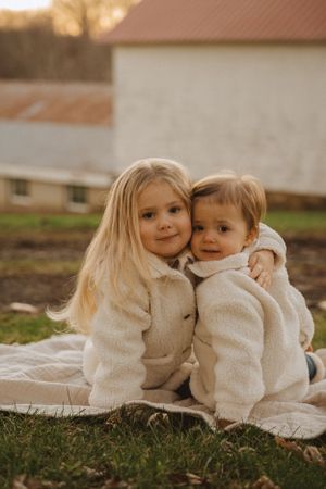 Two young children in cozy cream sweaters sitting together on blanket outdoors.