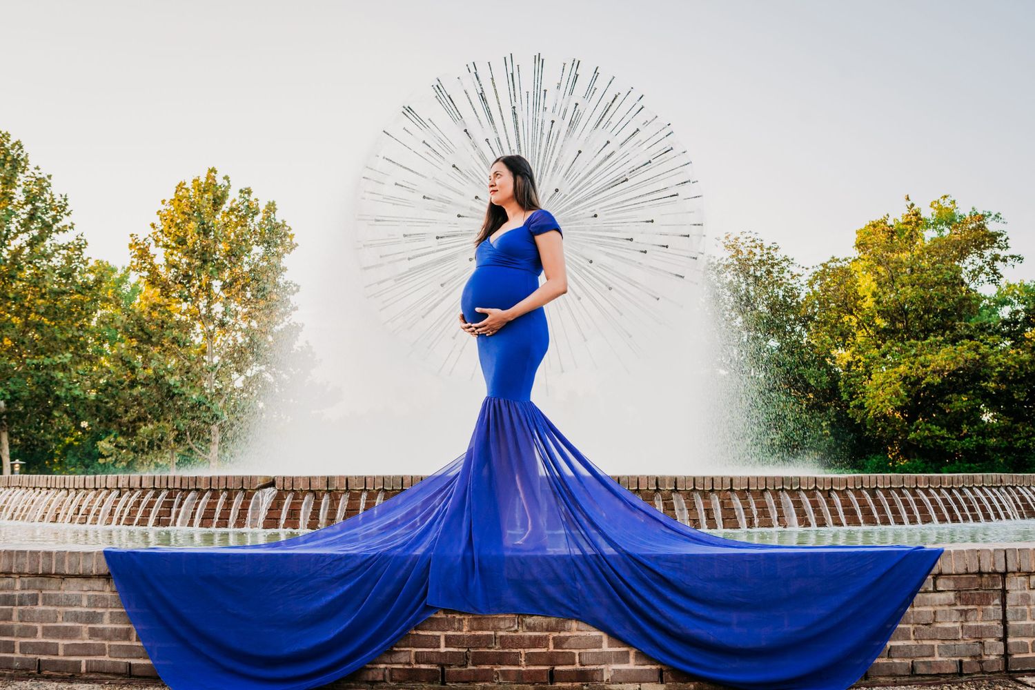 A dramatic maternity photo with a flowy royal blue gown in front of a sunburst fountain and autumn trees.
