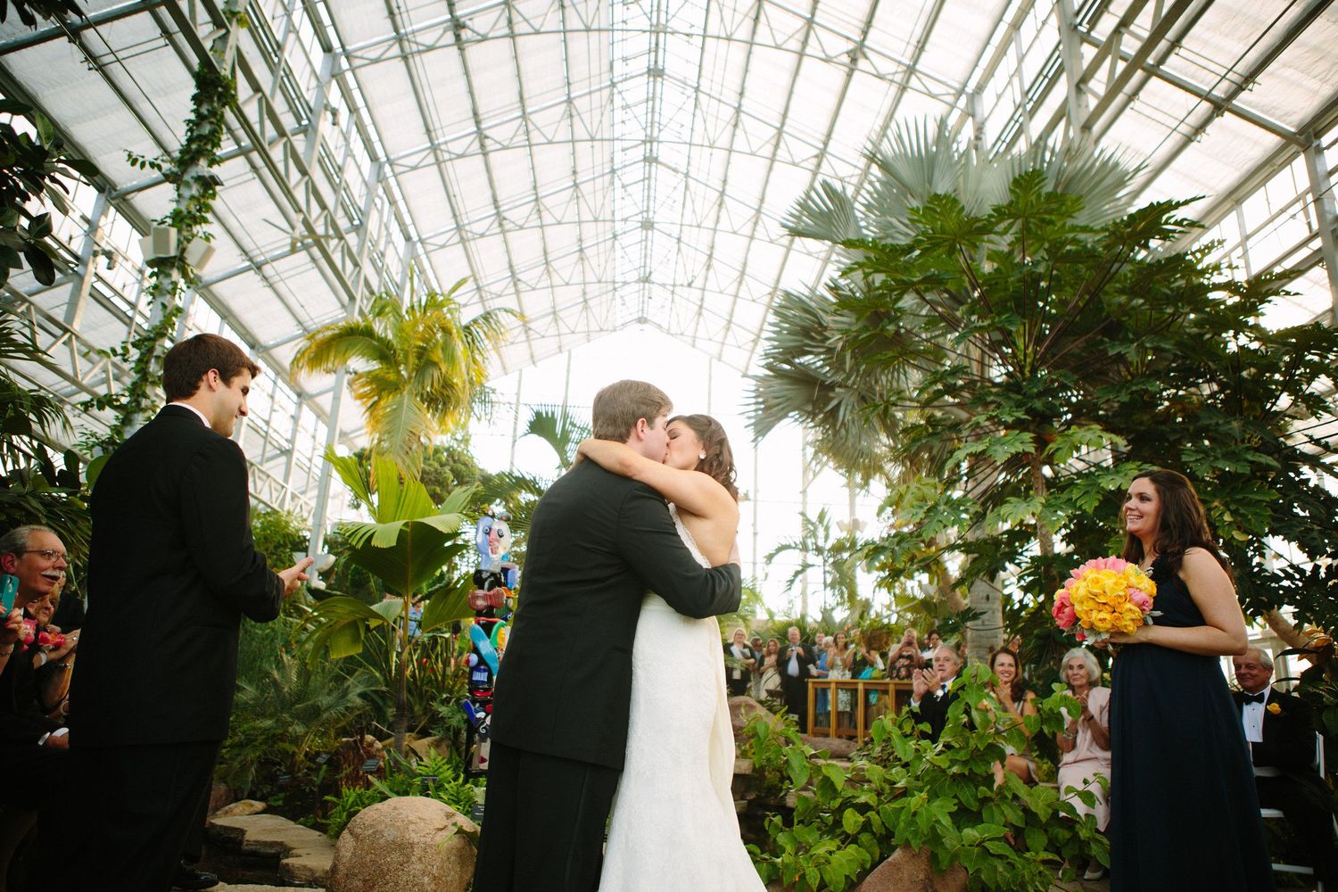 Greenhouse wedding ceremony with tropical plants and gathered guests.