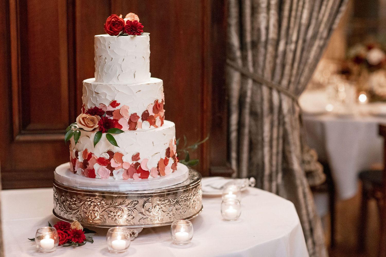 Three-tiered white wedding cake adorned with red and pink flowers on ornate silver stand.