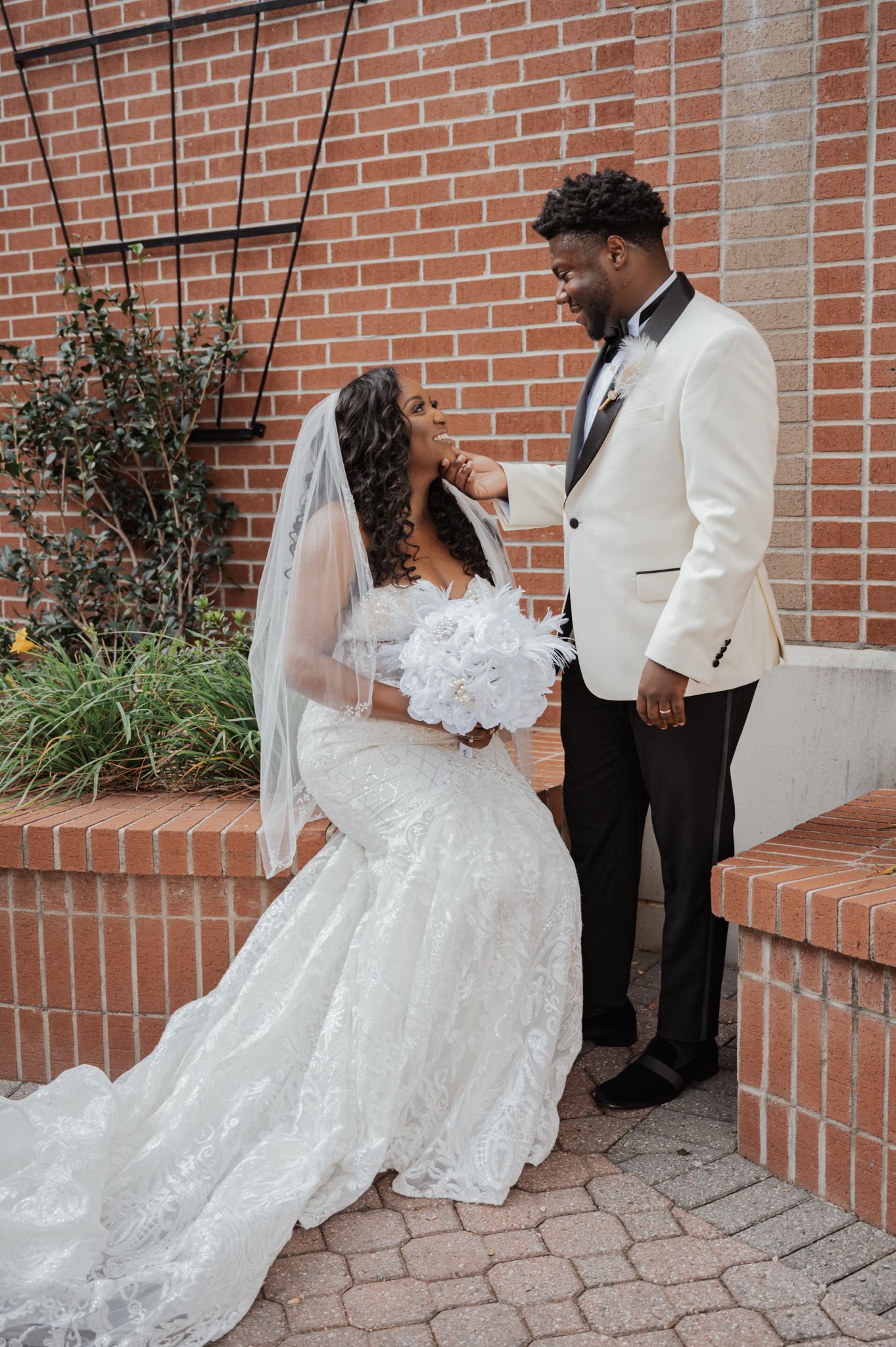 A couple in wedding attire poses against a brick wall with landscaping.