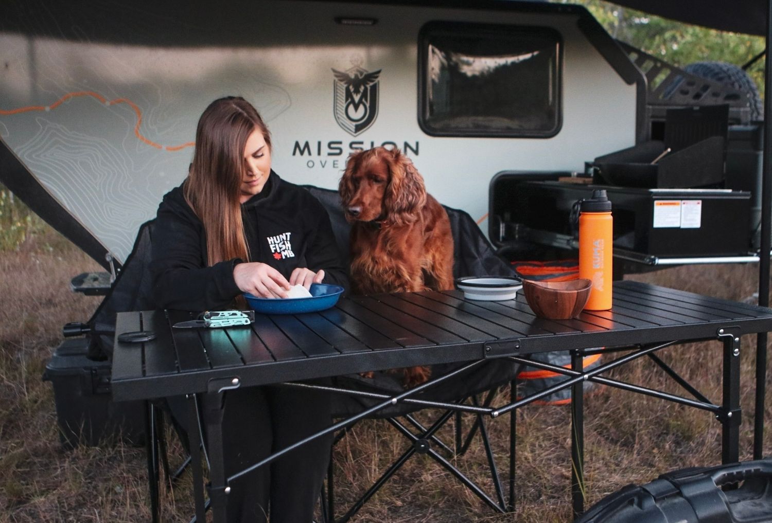 Camping setup with folding table and brown dog near travel trailer.