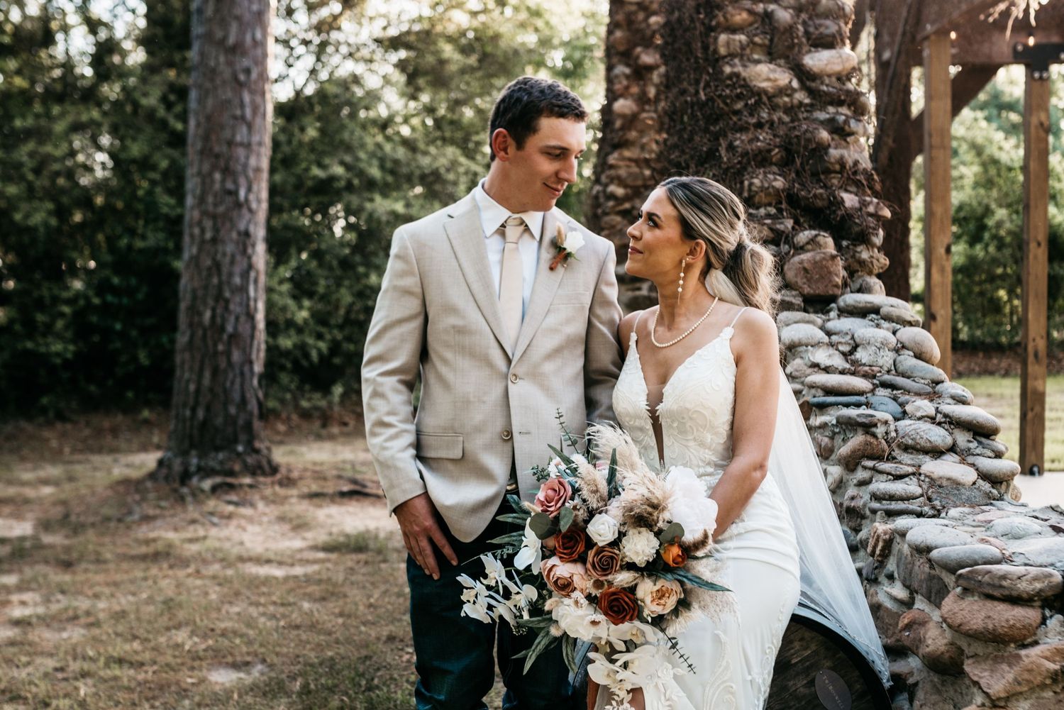Wedding couple poses outdoors with stone wall backdrop and lush floral bouquet.