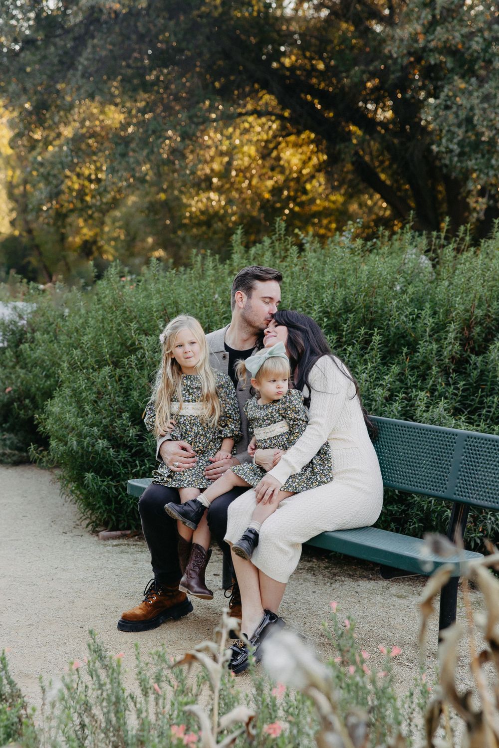 Intimate family portrait on a park bench with lush greenery and golden sunlight in background.