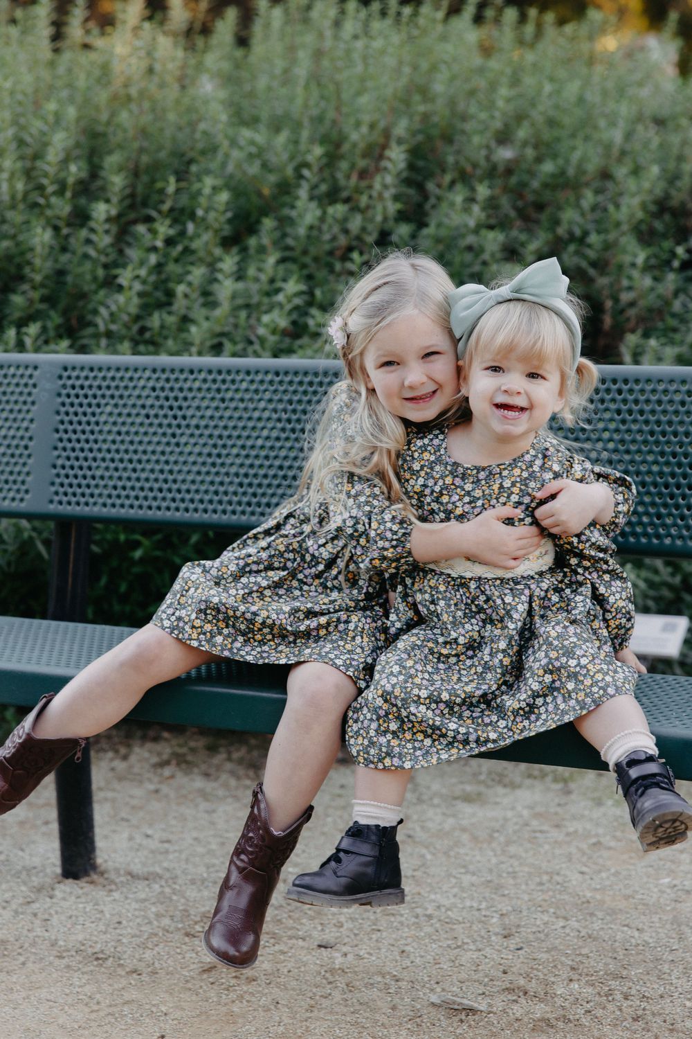 Two children in matching floral dresses sit together on a park bench surrounded by green foliage.