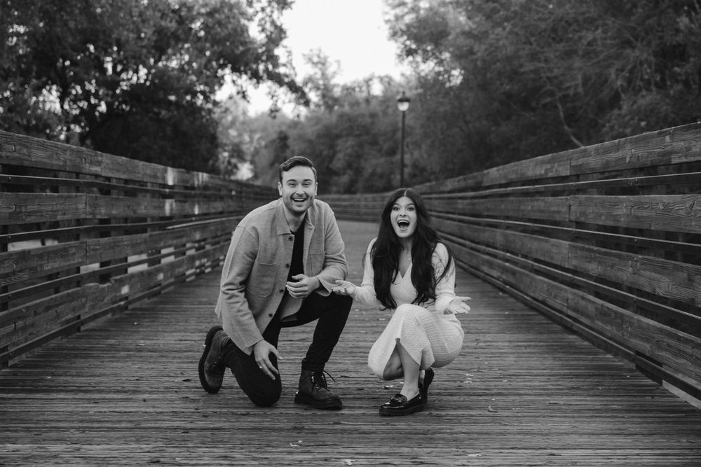 Black and white portrait of couple crouching on wooden bridge in park setting.