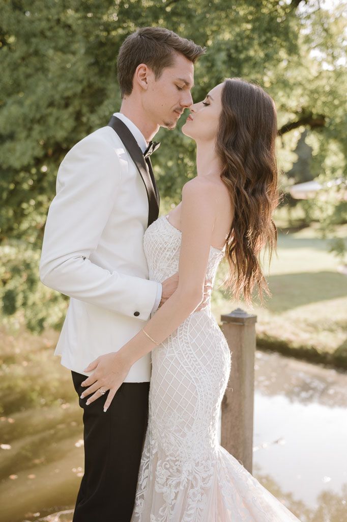 A romantic wedding couple embraces outdoors in elegant white attire with natural scenery in the background.