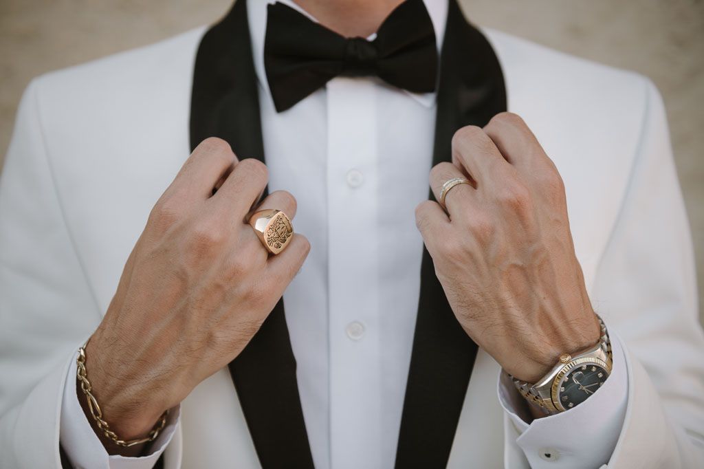 Detail shot of hands adjusting a black bowtie with luxury accessories and white tuxedo jacket.