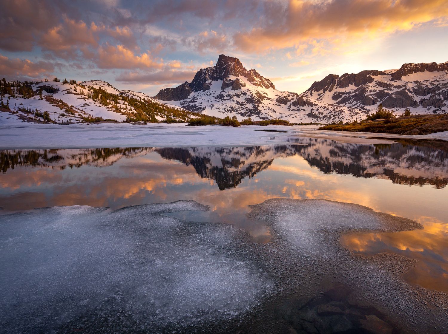 Snowy mountain peak reflects in partially frozen lake during dramatic sunset with orange clouds in winter landscape.