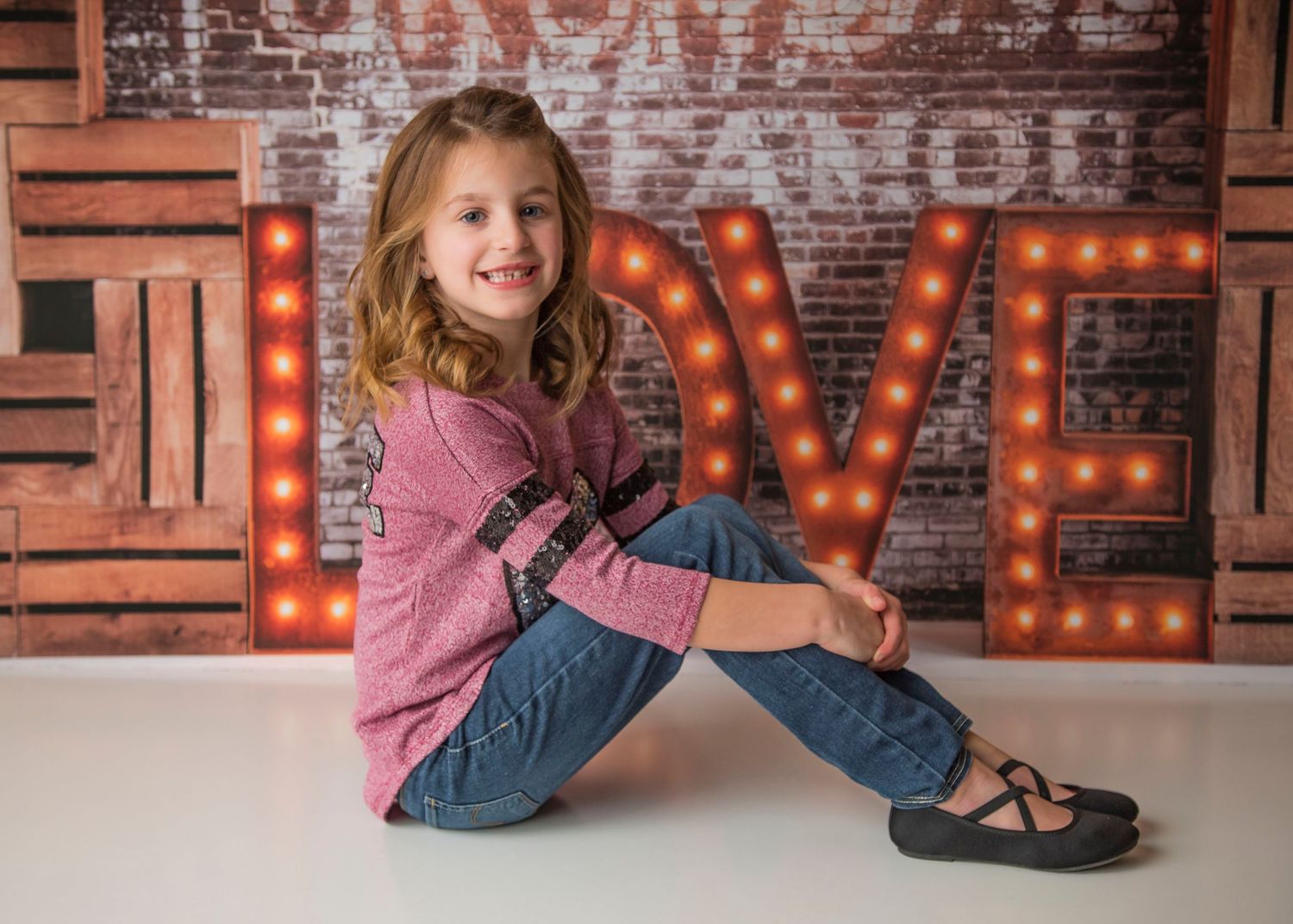 Child in pink top and jeans sits casually against rustic backdrop with glowing LOVE sign.