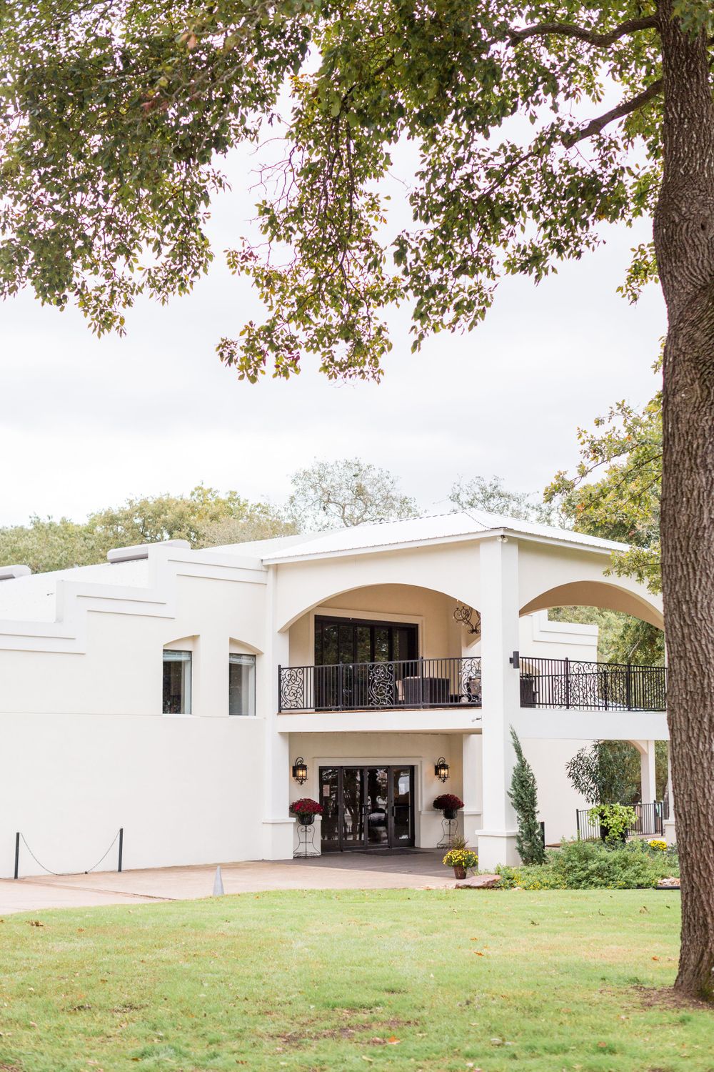 Elegant white two-story wedding venue with arched balcony and lush landscaping under mature oak trees.