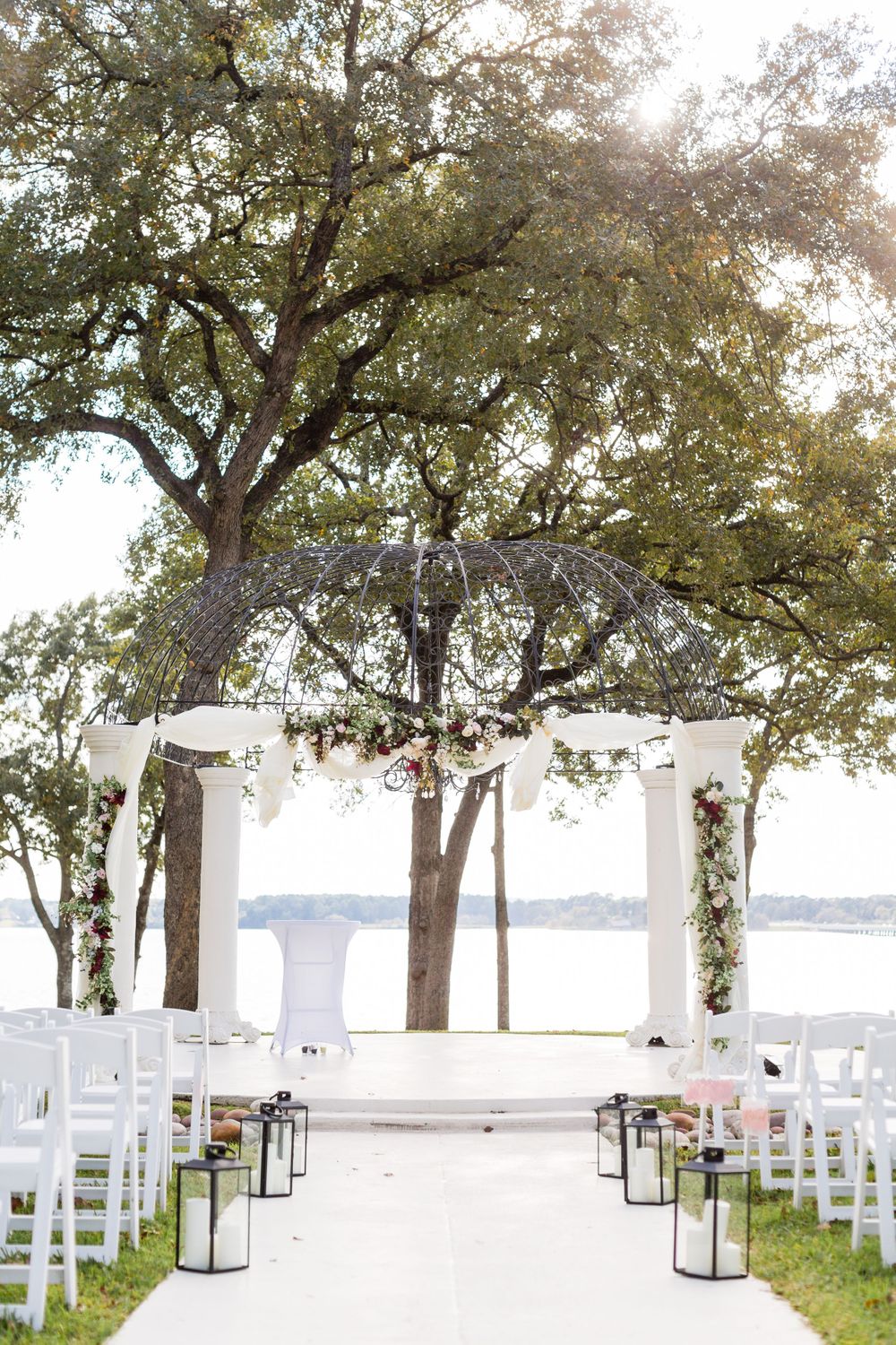 White wedding ceremony setup with lantern-lined aisle under oak trees beside water.
