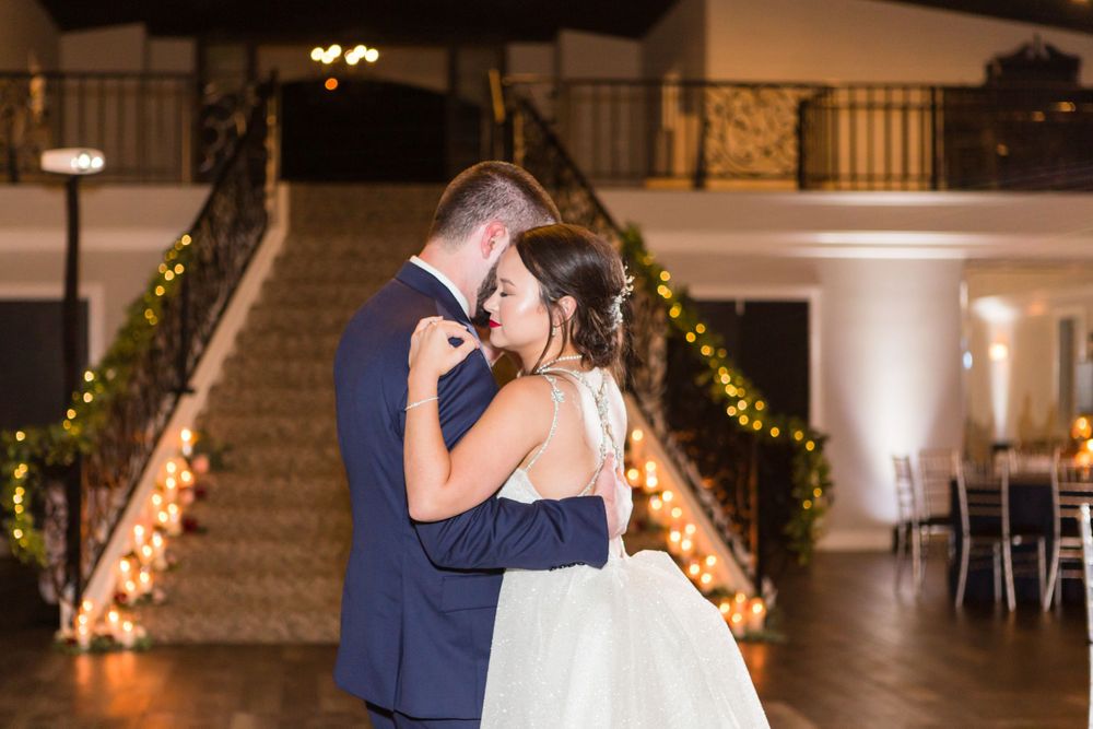 A couple shares their first dance at a wedding reception under warm lighting with decorated stairs in the background.